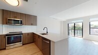 This single-floor unit showcases a contemporary design with a clean, open layout that connects the kitchen and living areas seamlessly. The kitchen is well-appointed with warm wood-tone cabinetry, white subway tile backsplash, a white quartz peninsula with an undermount sink and matte black faucet, and a full suite of stainless steel appliances including a range, microwave, and dishwasher. Light hardwood flooring extends throughout the main living areas, contributing to a bright and cohesive aesthetic.

The living area is spacious and sun-filled, anchored by expansive floor-to-ceiling sliding glass doors that open onto a private balcony with views of surrounding greenery, allowing abundant natural light to flood the interior. The neutral white walls and clean architectural lines enhance the airy atmosphere.

The bedroom is a comfortable, well-proportioned room featuring a large black-framed window that provides natural light and a closet for storage. The bathroom is finished with beige tile flooring, a tiled tub-shower surround, a wood-tone vanity, and a clean, functional layout. Throughout the unit, consistent finishes and a modern palette create a cohesive and polished living environment.