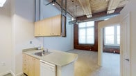 This single-floor loft showcases a compelling industrial aesthetic defined by a striking combination of exposed red brick walls, rustic wood-beam ceilings, and open ductwork that together create a warm yet raw architectural character. The soaring ceiling height amplifies the sense of openness throughout, while a ceiling fan with pendant lighting adds both function and visual interest overhead.

The kitchen area features light wood cabinetry mounted on adjustable shelving tracks, a laminate countertop with an integrated sink, and a dishwasher — providing practical functionality within the open-plan layout. The kitchen flows naturally into the main living area, where polished concrete-style flooring transitions seamlessly underfoot.

Large, multi-pane windows set within the exposed brick walls flood the interior with abundant natural light, offering bright, airy views to the outside. A secondary room, visible through a doorway, echoes the same brick-and-window aesthetic, maintaining a cohesive industrial character throughout the space.

The overall ambiance is loft-inspired and versatile, combining the charm of historic building materials with a flexible, open floor plan suited to a variety of living or working arrangements.