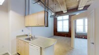 This single-floor loft showcases a compelling industrial aesthetic defined by a striking combination of exposed red brick walls, rustic wood-beam ceilings, and open ductwork that together create a warm yet raw architectural character. The soaring ceiling height amplifies the sense of openness throughout, while a ceiling fan with pendant lighting adds both function and visual interest overhead.

The kitchen area features light wood cabinetry mounted on adjustable shelving tracks, a laminate countertop with an integrated sink, and a dishwasher — providing practical functionality within the open-plan layout. The kitchen flows naturally into the main living area, where polished concrete-style flooring transitions seamlessly underfoot.

Large, multi-pane windows set within the exposed brick walls flood the interior with abundant natural light, offering bright, airy views to the outside. A secondary room, visible through a doorway, echoes the same brick-and-window aesthetic, maintaining a cohesive industrial character throughout the space.

The overall ambiance is loft-inspired and versatile, combining the charm of historic building materials with a flexible, open floor plan suited to a variety of living or working arrangements.