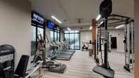 This fitness center is a comprehensively outfitted facility designed to support a wide range of workout routines. The space is organized into distinct zones that flow seamlessly into one another, offering an intuitive layout for users of all fitness levels. The cardio area lines one side of the room and features a row of treadmills, ellipticals, and a stationary spin bike, all positioned in front of expansive floor-to-ceiling windows that provide natural light and outdoor views, creating an energizing environment during workouts. Mounted flat-screen TVs are positioned throughout the space for entertainment.

The strength training area is centrally located and includes a multi-functional cable and power rack system with TRX suspension trainers, accommodating a variety of resistance and functional training exercises. A dedicated free weights section features a full rack of dumbbells, a weight bench, and a large mirrored wall, making it easy to monitor form during lifts. A heavy punching bag and a rack of medicine balls and kettlebells round out the functional fitness offerings.

The facility is finished with light wood-effect flooring, warm-toned accent columns, and ceiling fans paired with linear LED lighting, contributing to a clean and modern aesthetic throughout. The open, airy layout ensures ample room to move between equipment comfortably.