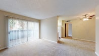 This single-story unit is designed with a practical, open layout that connects the main living areas seamlessly. The living room is generously sized, finished with wall-to-wall carpet in a neutral beige tone, and features a textured ceiling. A full-width sliding glass door fitted with vertical blinds fills the space with natural light and provides direct access to an outdoor patio, offering views of surrounding greenery.

Adjacent to the living area, the kitchen is separated by a partial dividing wall, creating a semi-open feel. It is equipped with white cabinetry, a white electric range, a refrigerator, and light-colored countertops. The kitchen floor transitions to wood-style vinyl, distinguishing it from the carpeted living spaces.

The bedroom is a well-proportioned room with plush carpet, a ceiling fan with light fixture, a dedicated closet, and a window that admits natural light. Warm beige walls are consistent throughout the unit, contributing to a cohesive, neutral aesthetic.

The bathroom includes a full tub and shower combination with a white surround, a toilet, and a vanity with a light countertop. Wood-style flooring carries through this space, complementing the clean, white fixtures. Overall, the unit presents a functional and straightforward residential layout suited for everyday living.