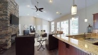 This single-floor residence in Jefferson offers a thoughtfully designed open-concept layout that seamlessly connects the living and kitchen areas. The soaring vaulted ceilings create an impressive sense of height and airiness throughout the main living space, further enhanced by recessed lighting and a ceiling fan that contributes both style and comfort. The kitchen features a substantial island or bar-height counter finished in warm granite, complemented by rich cherry-toned wood cabinetry and elegant pendant lighting overhead. The living area is anchored by dark hardwood-style tile flooring and furnished with deep chocolate-brown leather sofas arranged around a glass-top coffee table, lending the space a sophisticated yet welcoming ambiance. Neutral warm beige tones on the walls provide a cohesive backdrop throughout. Large windows along the rear wall flood the interior with generous natural light and offer views of the surrounding greenery outside. Decorative touches such as a classic starburst wall accent and floral arrangements add character to the space. The overall design strikes a balance between open, airy architecture and warm, inviting finishes, making it an ideal space for both relaxing and entertaining.