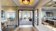 The Signal Hill Club Room in Manassas, Virginia is a thoughtfully designed community gathering space that blends sophistication with comfort across a single expansive floor. The entrance greets visitors with a stylish foyer featuring a coffered tray ceiling with warm accent lighting, decorative pendant fixtures, and framed artwork, setting an upscale tone from the moment of arrival. Large wood-framed doorways connect the spaces seamlessly, creating an open and inviting flow throughout. The main lounge area is anchored by generous seating arrangements, including plush sectional sofas, accent chairs in warm gold and cool grey tones, and patterned area rugs that add texture and depth. Tray ceilings with ambient lighting and striking orb pendant light fixtures lend architectural interest overhead, while floor-to-ceiling windows flood the space with abundant natural light and offer direct views of the outdoor pool area and surrounding community. A dedicated kitchen features stainless steel appliances, flat-panel cabinetry, a marble-topped island with navy tile detailing, globe pendant lights, and bar seating — ideal for hosting and entertaining. Throughout the club room, neutral wall tones, carefully curated décor, and patterned carpeting create a warm, polished atmosphere well-suited for both social gatherings and quiet relaxation.