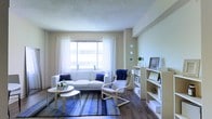 This single-floor living space presents a well-organized and inviting layout centered around a comfortable living area. The room features dark hardwood flooring that grounds the space, contrasting elegantly with the light, neutral-toned walls finished with a textured ceiling.

A large window dressed with sheer white patterned curtains allows natural light to flood the room, creating a bright and airy atmosphere. The living area is anchored by a white sofa accented with navy blue throw pillows and a coordinating blue striped area rug, establishing a cohesive blue and white design theme throughout.

A natural wood-framed accent chair adds warmth and a contemporary touch beside the sofa. A set of round nesting coffee tables in a light wood finish sits at the centre, complementing the overall aesthetic. Along the wall, a white bookcase and a cube storage unit provide practical display and storage solutions, decorated with framed artwork and small ornamental pieces. Circular wall mirrors add a decorative focal point, contributing to the room's polished and thoughtfully curated ambiance.