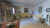 This single-floor space presents a comfortable and traditionally styled living area designed for relaxed everyday living. The living room serves as the central gathering space, featuring plush wall-to-wall carpeting in a soft neutral tone that contributes to the room's warm ambiance. The seating arrangement includes a yellow upholstered sofa adorned with floral accent pillows, a cream-colored armchair, and a striped wingback chair, all centered around a decorative wooden coffee table. Two matching table lamps with classic bases provide warm ambient lighting, complementing the natural light that filters through a window dressed with red-toned curtains on the far wall. Framed artwork on the walls adds a finishing touch to the décor. A wooden cabinet or hutch is visible near the window, offering additional storage and display space. In the foreground, a dining area is partially visible, suggesting a seamlessly connected layout between the living and dining spaces. The overall atmosphere is inviting, with a cohesive traditional design style maintained throughout the visible areas of the home.
