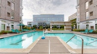 Located at 2838 Fremont Ave S in the Uptown neighborhood of Minneapolis, Minnesota, this outdoor amenity space serves as a vibrant communal hub nestled within a modern multi-story residential building. The centerpiece of the courtyard is a generously sized outdoor swimming pool with clear turquoise water, complete with dedicated entry steps, safety railings, and a surrounding pool deck outfitted with teal lounge chairs positioned for relaxation and sun exposure. Adjacent to the main pool sits a raised spa or hot tub enclosure, providing a distinct zone for hydrotherapy and unwinding. The pool area is enclosed by a sleek iron fence and offers open views of the surrounding urban skyline, including a striking glass-facade commercial building that forms a dramatic backdrop. Flanking the pool deck is a spacious paved courtyard designed for social gatherings, featuring high-top bar tables and chairs shaded by large umbrellas in both teal and navy tones. Mature trees rise from curated landscaped planting beds, adding greenery and a sense of natural enclosure to the space. The hardscape incorporates alternating stone and concrete pavers in a structured geometric pattern. Architectural columns with a distinctive striped brown-and-tan cladding anchor the space visually, while glass-fronted building entrances provide seamless indoor-outdoor connectivity. The overall ambiance is polished, modern, and well-suited for both active recreation and casual outdoor socializing.