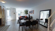 This well-appointed two-floor space combines contemporary design with a warm, livable aesthetic. The main living and dining area is open-plan, creating a seamless flow between spaces. Light hardwood flooring runs throughout, contributing to a bright and airy atmosphere. A dining area anchors the foreground with a dark walnut table surrounded by sleek upholstered chairs, complemented by a textured area rug underneath. A large statement mirror leans against one wall, adding depth and dimension to the room. The living area beyond features a dark leather sofa accented with vibrant orange throw pillows, grounding the space with contrast. A striking fiddle-leaf fig tree adds a lively natural element, drawing the eye toward the expansive floor-to-ceiling windows at the rear. These windows flood the space with natural light and offer views of lush greenery outside, seamlessly connecting the interior to the outdoors. Tasteful wall art and subtle decorative accents, including a mounted deer head and framed artwork, give the space a curated, modern character. The clean white walls and smooth ceilings keep the overall palette fresh and neutral, allowing the furnishings and natural light to take center stage. The second floor adds additional living versatility to this thoughtfully designed home.