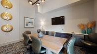 This conference room offers a polished and functional environment ideal for meetings and collaborative work. The space is defined by notably tall ceilings with an elevated tray detail, lending an open, airy quality that sets it apart from a typical meeting room. A central light wood rectangular table is surrounded by multiple ergonomic leather chairs, comfortably accommodating a group of six to eight participants.

A large flat-screen display is wall-mounted prominently, serving as the focal point for presentations. Beneath it, a continuous run of dark-toned built-in cabinetry with a clean white countertop provides ample storage and workspace along one wall. A table lamp with a gold base adds warm, ambient lighting to complement the modern multi-bulb pendant chandelier overhead.

Decorative elements elevate the room's aesthetic, including a series of three hammered gold disc wall sculptures, framed abstract artwork in warm tones, and tasteful ceramic and botanical accents. A patterned carpet in earth tones grounds the space with texture and visual interest. A glazed door allows natural light to filter in, contributing to the room's bright, professional, and welcoming atmosphere.