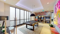 The Games Room at 200 Redpath is a thoughtfully designed amenity space located in Toronto's Midtown neighbourhood, offering a diverse range of entertainment and social features across a single floor. The space is anchored by a sleek pool table positioned beneath a bold dome pendant light, set against floor-to-ceiling windows that frame expansive city views. Adjacent to the pool table, a collection of classic arcade and pinball machines lines a vibrant comic-style mural wall, adding a playful, energetic character to the room. A dedicated bowling alley with integrated scoring displays and ambient LED lighting provides an additional recreational focal point, accompanied by a clean white kitchenette with a textured chevron accent wall nearby. Throughout the space, light-toned herringbone hardwood flooring lends warmth and continuity. A lounge area features a double-height volume with floor-to-ceiling windows, a glass-railed staircase, large-scale artwork, and lush indoor plants, creating an airy, sophisticated atmosphere. A separate seating nook with a large city-view window offers a quieter retreat. The kitchenette area is highlighted by a striking diamond-patterned copper and white tile backsplash, a wine fridge, and a marble dining table. The overall design balances contemporary elegance with bold, playful elements throughout.