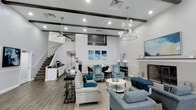This stylish communal lounge space is designed to impress from the moment you enter. The soaring ceilings are accented by rich dark wood exposed beams that add warmth and architectural character to the otherwise crisp, white interior. Recessed lighting and a sophisticated chandelier illuminate the space, complementing the natural light that filters throughout. Dark hardwood-style flooring anchors the room, providing an elegant contrast to the light-toned walls and furnishings. The seating area is thoughtfully arranged with a plush light gray sofa, coordinating blue accent chairs, and a round white coffee table, all tied together by a neutral area rug. A fireplace with a refined tile surround serves as a focal point along one wall, topped by a large flat-screen display above the mantle. Coastal-inspired artwork in shades of blue adds a cohesive design theme throughout. A beverage and refreshment station is conveniently tucked along one side, ideal for gatherings. A staircase with dark wood treads adds visual depth and architectural interest to the rear of the space. Overall, the lounge exudes a polished yet inviting ambiance suited for relaxation and community engagement.