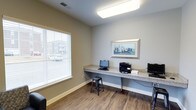 This single-floor space is designed as a functional business center, thoughtfully laid out to accommodate productivity needs. The room features a long, built-in granite-topped workstation that runs along one wall, providing ample desk space for multiple users simultaneously. The workstation is equipped with desktop computers, a multifunction printer, and a laptop, making it well-suited for everyday computing and printing tasks.

The flooring is hardwood-style, lending a clean and polished appearance to the space. Neutral-toned walls in soft beige and gray create a calm, professional atmosphere, complemented by white baseboards that add a crisp finishing detail. A large window fitted with white horizontal blinds allows natural light to fill the room while offering a view of the surrounding exterior. Overhead, a flush-mounted fluorescent light fixture ensures consistent, bright illumination throughout.

A framed cityscape artwork is displayed above the workstation, adding a subtle decorative touch to the otherwise utilitarian setting. A simple metal stool is positioned at the workstation, offering seating for users. Overall, the space is clean, efficiently organized, and well-equipped for business and administrative tasks.