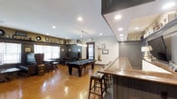 This single-floor entertainment and lounge space is thoughtfully designed to offer a rich, social atmosphere with a classic, upscale feel. At its centerpiece sits a full-sized billiards table set atop polished concrete floors that reflect warm ambient light throughout the room. A built-in bar lines one side of the space, complete with a wooden countertop and leather-upholstered bar stools, making it an ideal spot for casual conversation and drinks. The interior carries a cohesive dark wood aesthetic, with wainscoting panels, dark-trimmed accents, and warm recessed lighting that create an inviting, pub-like ambiance. A decorative pendant light fixture adds a focal point above the central gathering area. Along the perimeter, framed artwork and decorative items are displayed on shelving, lending a personalized character to the space. A cozy lounge nook with tufted booth-style seating and small café tables provides a more intimate area for guests. Natural light filters in through window blinds, complementing the interior lighting. This space is well-suited for recreational use, private events, or community gatherings, offering a seamless blend of functionality and sophisticated design throughout its open, single-level layout.