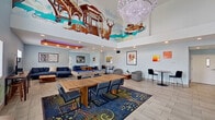 This single-story community clubhouse is defined by its dramatic double-height ceilings and an open, versatile layout designed to encourage social interaction and recreation. The entrance lobby welcomes visitors with floor-to-ceiling glass doors, clerestory windows that flood the space with natural light, and light-toned tile flooring that runs throughout. Workstations near the entry provide a practical area for administrative use.

The main gathering area is the visual centerpiece of the clubhouse, anchored by a large, colorful mural spanning the upper walls and depicting wildlife and architectural scenes. A grand crystal chandelier hangs overhead, adding an elegant contrast to the playful artwork. Recessed lighting complements the ambient glow throughout.

Furnishings are arranged to support multiple activities simultaneously. A pool table and foosball table cater to recreational use, while a substantial wooden dining table surrounded by navy upholstered chairs serves as a collaborative hub. Multiple seating clusters, featuring grey accent chairs, navy sofas, and bistro-height tables, offer flexible spaces for conversation or quiet relaxation.

Framed artwork is displayed throughout, adding layers of visual interest. The overall atmosphere is lively yet welcoming, balancing energetic recreational elements with comfortable, well-appointed lounge spaces.