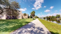 This single-floor property is situated within a well-maintained residential community that evokes a distinctly tropical Florida atmosphere. The exterior environment is one of the space's most defining features, showcasing a paved pedestrian pathway that winds alongside a calm canal, framed by tall, mature palm trees and broad shade trees that cast dappled light across the walkway. Lush, well-kept green lawns border both sides of the path, contributing to the community's clean and serene character. A classic lamppost stands along the walkway, adding a timeless aesthetic detail to the outdoor corridor. The waterway running parallel to the path reflects the open sky and surrounding greenery, creating a peaceful natural backdrop. Residential buildings with a neutral, earth-toned exterior are visible along the route, blending harmoniously with the landscape. The overall setting conveys a strong sense of outdoor livability, with an emphasis on green space, water access, and walkable surroundings. The interior images captured were too dark to yield detailed information, though the property occupies a single floor, suggesting a compact and accessible layout. The surrounding community infrastructure points to a well-established neighborhood designed with comfort and outdoor enjoyment in mind.
