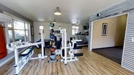 This single-floor fitness center is thoughtfully designed to support a comprehensive workout experience. The space features light hardwood flooring throughout, creating a clean and welcoming environment that contrasts with the neutral white walls. Industrial-style pendant lights hang from the ceiling, providing warm, even illumination across the room.

At the center of the space stands a multi-station strength training machine, ideal for a variety of resistance exercises. A dedicated medicine ball rack holds an assortment of weighted balls in multiple sizes, catering to functional fitness and core training needs. A full-length mirror along one wall enhances the sense of openness while allowing users to monitor their form during workouts.

The cardio section, partially separated by a decorative stone or brick accent wall, houses treadmills positioned to maximize the use of available space. A mounted flat-screen television is visible in this area, providing entertainment during cardio sessions. Natural light filters in through multiple windows, giving the space an airy, energizing feel.

The layout is practical and efficient, allowing users to move freely between equipment zones. Framed artwork on the walls and tasteful architectural details add a polished touch, making this fitness center both functional and visually appealing.