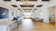 This single-story space presents a refined coastal aesthetic throughout its open, well-appointed interior. The reception area serves as the visual centerpiece, featuring a bold blue subway-tiled front desk complemented by a geometric, blue-and-white patterned accent wall. Coastal-themed artwork, including a prominent heron print, reinforces the seaside design concept.

The coffered ceiling is a standout architectural element, with dark wood-paneled insets framed by crisp white beams and illuminated by two elegant multi-arm chandeliers. Light wood-look porcelain tile flooring extends throughout, creating a seamless, airy continuity between spaces.

The main lounge area is generously proportioned, furnished with natural wood-framed seating and neutral upholstery arranged to encourage conversation. Floor-to-ceiling windows and glass doors frame unobstructed views of an outdoor pool and palm trees, flooding the interior with abundant natural light.

A secondary lounge area features additional seating, a dark wood coffered ceiling detail, and direct access to the pool deck. A separate service area includes white cabinetry with granite countertops. Together, these spaces convey a bright, welcoming, and resort-inspired ambiance throughout.
