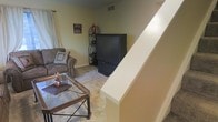 This two-story space is characterized by its practical, comfortable layout connecting multiple levels via an interior staircase. The living area, visible from the staircase landing, features wall-to-wall carpeting in neutral tones that complement the warm beige walls throughout. The space accommodates a traditional seating arrangement, including an upholstered sofa accented with decorative pillows, paired with a glass-top coffee table that adds a touch of lightness to the room. A large television serves as a focal point, flanked by a decorative wrought-iron shelving unit that provides both storage and visual interest. Natural light filters into the living area through curtained windows, contributing to the room's inviting atmosphere. Ceiling vents indicate a central HVAC system for year-round comfort. The staircase, finished with a clean white newel post, provides a clear visual division between the living space and the upper level. The overall ambiance is warm and functional, with decor choices that emphasize a lived-in, homey character throughout the main living area.