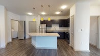 This single-floor residence showcases a thoughtfully designed open-concept layout, with the kitchen serving as a central highlight. Dark espresso cabinetry lines the walls, contrasting elegantly with light granite countertops that extend across ample workspace. Stainless steel appliances — including a refrigerator, range, microwave, and dishwasher — provide a polished, modern finish. A freestanding center island with a matching granite surface offers additional prep space and casual seating, while three yellow pendant lights hang above, adding a distinctive accent. Wood-look plank flooring flows throughout the main living areas, lending warmth and continuity.

The bathroom mirrors the kitchen's aesthetic, featuring dark cabinetry with granite countertop, a full tub-shower combination with tile surround, and wood-look flooring. A dedicated laundry room houses a full-size side-by-side washer and dryer along with wire shelving above for storage convenience.

The bedroom is bright and airy, benefiting from multiple windows that invite natural light and views of the surrounding exterior. A ceiling fan provides comfort, and carpet underfoot adds softness. A generously sized walk-in closet with multi-tiered wire shelving provides extensive storage capacity, completing this well-appointed space.