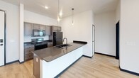 Located at 612 6th St in Rapid City, South Dakota, this 2-bedroom, 2-bathroom unit offers a well-designed single-level layout with modern finishes throughout. Light wood-tone flooring flows seamlessly across the entire space, complemented by crisp white walls and bold dark baseboards that give the interiors a clean, contemporary edge.

The kitchen serves as a central highlight, featuring an expansive peninsula island with dark granite-style countertops, an undermount sink with a matte black faucet, and stylish pendant lighting overhead. Taupe-toned cabinetry lines the walls alongside a full suite of stainless and black appliances, including a refrigerator, range, and microwave.

Both bedrooms are well-proportioned and bathed in natural light through large sliding windows that frame elevated views of the Rapid City skyline and surrounding landscape. The primary bedroom includes a built-in double-door closet, while exposed ductwork and ceiling fans add an industrial character to the rooms.

The two bathrooms are finished consistently with dark-stained vanity cabinets, dark countertops, large mirrors, and warm vanity lighting. One bathroom features a tub-shower combination. Additional spaces include a utility room and a dedicated closet, rounding out the functional layout of this modern urban residence.
