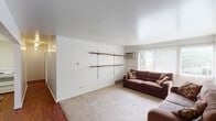 Located at 208 Cedar St in East Lansing, Michigan, this single-floor unit offers a practical and functional layout suited for comfortable everyday living. The living area is a welcoming space featuring light-toned carpet, white vertical panel wainscoting along one wall, and built-in wooden shelving that adds a touch of character. A wall-mounted air conditioning unit and baseboard heating provide year-round climate control, while large windows dressed with vertical blinds allow generous natural light to fill the room and offer views of the surrounding greenery outdoors. The bedrooms are carpeted and bright, each featuring large windows with vertical blinds and baseboard heating, creating a clean, airy atmosphere. The bathroom is well-appointed with a dual-sink vanity set against dark cabinetry and a wide mirror above, complemented by a full tub and shower combination with tiled walls. Hardwood flooring accents the entryway and bathroom areas, providing a pleasant contrast to the soft carpet found throughout the rest of the unit. The overall layout, as seen from the dollhouse view, is open and efficiently organized, with a kitchen, ceiling fan, and connected common areas making the space both practical and inviting for residents.