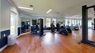 This single-floor fitness center offers a comprehensive workout environment designed to accommodate a range of exercise needs. The space is finished with warm hardwood flooring throughout, providing a clean and durable surface well-suited for fitness use. The walls feature a two-tone paint scheme, with a deep navy blue on the lower half and a soft gray-blue on the upper half, creating a structured, energetic atmosphere.

Natural light fills the room through numerous white-trimmed windows positioned along multiple walls, offering views of the surrounding outdoor greenery. Large floor-to-ceiling mirrors are strategically placed to visually expand the space and allow users to monitor their form during exercise.

The fitness center is equipped with a variety of machines, including a treadmill, elliptical trainers, a multi-station cable weight machine, a leg press and rowing machine combo, and a recumbent bike. A medicine ball and additional free accessories complement the equipment selection. Overhead fluorescent lighting supplements natural light, ensuring consistent illumination throughout the day. Posted fitness guidelines are displayed on the wall, and a security camera is visible, reflecting attention to safety and facility management.