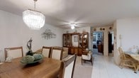 This single-floor residence offers a thoughtfully arranged and warmly decorated interior that blends classic style with comfortable living. The combined dining and living area serves as the heart of the home, where an eye-catching crystal chandelier anchors the dining space, casting a refined elegance over the wooden dining table and cushioned chairs. Light tile flooring runs throughout, lending a clean and cohesive feel to the space. A ceiling fan with a built-in light fixture keeps the living area comfortable, which is furnished with a classic wooden display cabinet showcasing a curated collection of decorative figurines and keepsakes. A wooden armchair and neutral-toned sofa add to the relaxed, homey atmosphere. The walls are adorned with tasteful metal wall art, contributing to the space's warm, personalized character. A hallway visible from the main living area leads to additional rooms, hinting at further living spaces beyond. The overall ambiance is one of a well-loved and carefully maintained home, rich in character, with classic furnishings and decorative touches that create an inviting and lived-in atmosphere throughout.