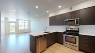This contemporary single-floor unit showcases a clean, modern design throughout, highlighted by light hardwood flooring and recessed lighting that create a bright, welcoming atmosphere. The open-concept kitchen is a standout feature, equipped with sleek dark-finish cabinetry that extends to the ceiling for ample storage, crisp white countertops, and a full suite of stainless steel appliances including a gas range, built-in microwave, dishwasher, and refrigerator. A peninsula with a double sink extends into the living area, making the kitchen ideal for both cooking and casual dining. The adjacent living area is generously proportioned, bathed in natural light from a large window that offers views of the surrounding neighborhood. The bedroom is a serene, well-sized retreat featuring the same warm hardwood flooring and a wall of built-in, floor-to-ceiling white cabinetry providing extensive closet and storage space. The bathroom presents a functional and contemporary layout with large-format tile flooring, dark-finish vanity cabinetry, a glass-enclosed shower, and a stacked washer/dryer unit conveniently integrated into the space. The overall design is cohesive and polished, combining practicality with a refined modern aesthetic.