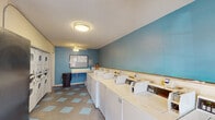 This shared laundry facility in Lafayette, Louisiana, is a well-equipped, single-floor space designed for practical, everyday use. The room is finished with light blue and teal accent walls that lend a clean, welcoming atmosphere, complemented by a textured ceiling and ceiling fan fixtures that provide ambient lighting and air circulation throughout.

The layout is efficiently organized, with a row of top-load washing machines lining one wall and stacked dryer units positioned along an adjacent wall, offering ample capacity for multiple users. Each machine features digital card-reader payment panels, providing a convenient, cashless laundry experience. A wall-mounted folding counter offers a dedicated surface for sorting and folding laundry, while a cork bulletin board nearby displays posted notices and facility information.

A snack and beverage vending machine is positioned near the entrance, adding a convenient amenity for residents during laundry sessions. The floor features a neutral tile with a teal checkerboard pattern, tying together the room's color scheme. A window along one wall allows natural light to enter, brightening the space. The entry door is equipped with a keypad access system, ensuring security for authorized users. Overall, the facility is functional, organized, and thoughtfully appointed.