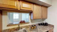This single-floor residence is designed with practicality and comfort in mind. The kitchen is a standout feature, outfitted with warm honey-toned wood cabinetry offering ample upper and lower storage, granite-look countertops, and a full suite of black appliances including a gas range, built-in microwave, refrigerator, and dishwasher. A pass-through window connects the kitchen to the adjacent living area, enhancing the sense of openness between spaces. The kitchen flooring is hardwood-style, adding a clean, durable finish underfoot.

The living and bedroom areas are carpeted in neutral tones, providing a soft, comfortable feel throughout. Each room is equipped with a ceiling fan with integrated lighting, contributing to both air circulation and ambient light. Sliding glass doors in one room open to an outdoor view, allowing natural light to fill the space, while additional windows with blinds provide adjustable brightness in other rooms.

The bathroom features tile flooring, a vanity with warm wood cabinetry, and a clean, straightforward layout with neutral wall tones. The entry area transitions from hard flooring into the carpeted living space, establishing a clear boundary between zones. Overall, the unit presents a cohesive, functional layout with consistent warm tones and practical finishes throughout.