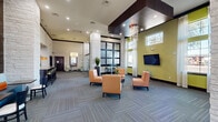 This generously proportioned single-floor amenity space is designed to accommodate both social gatherings and quiet relaxation. The main lounge area commands attention with its soaring double-height ceilings, accented by a dramatic dark wood coffered ceiling detail and an elegant chandelier. Chartreuse green accent walls, patterned carpet tiles, and large windows along the perimeter flood the space with natural light and views of the surrounding landscape. Comfortable lounge seating, including upholstered armchairs arranged around a central coffee table, creates an inviting social hub, while wall-mounted media displays add a modern, functional touch. Structural columns clad in stacked white stone add architectural character and visually anchor the open floor plan. A semi-private dining nook features tufted banquette seating in a vibrant teal, complemented by accent artwork. The fully equipped kitchen is appointed with rich dark wood cabinetry extending floor to ceiling, granite countertops, a mosaic tile backsplash, stainless steel appliances, and a kitchen island with bar seating — ideal for catering or community events. The seamless transition between tiled and carpeted flooring zones helps define functional areas within the open layout, creating a cohesive and welcoming environment throughout.