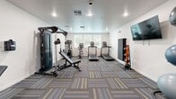 Located at 771 Harrington Way in Folsom, California, this well-appointed fitness center offers a comprehensive workout environment within a efficiently utilized single-floor layout. The room features crisp white walls, recessed lighting, and stylish patterned carpet tiles in gray, blue, and yellow tones that lend a modern, energetic feel to the space.

The cardio zone is equipped with multiple treadmills and an elliptical trainer, positioned near windows fitted with horizontal blinds that allow natural light to filter in. A Matrix multi-station cable machine and an adjustable weight bench anchor the strength-training area, providing versatile options for resistance workouts. A dedicated leg press machine occupies its own corner, accompanied by a window that adds additional brightness to the area.

On the opposite wall, a wall-mounted flat-screen TV serves as a focal point above a well-stocked free weights section, featuring a full dumbbell rack, kettlebells, and stability balls in various sizes. Medicine ball storage rounds out the functional training offerings. The room is accessed through glass-paneled doors, offering visibility into the space and contributing to an open, welcoming atmosphere. Ceiling-mounted recessed lights ensure consistent, even illumination throughout, making this fitness center a practical and inviting facility for daily exercise.