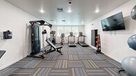 Located at 771 Harrington Way in Folsom, California, this well-appointed fitness center offers a comprehensive workout environment within a efficiently utilized single-floor layout. The room features crisp white walls, recessed lighting, and stylish patterned carpet tiles in gray, blue, and yellow tones that lend a modern, energetic feel to the space.

The cardio zone is equipped with multiple treadmills and an elliptical trainer, positioned near windows fitted with horizontal blinds that allow natural light to filter in. A Matrix multi-station cable machine and an adjustable weight bench anchor the strength-training area, providing versatile options for resistance workouts. A dedicated leg press machine occupies its own corner, accompanied by a window that adds additional brightness to the area.

On the opposite wall, a wall-mounted flat-screen TV serves as a focal point above a well-stocked free weights section, featuring a full dumbbell rack, kettlebells, and stability balls in various sizes. Medicine ball storage rounds out the functional training offerings. The room is accessed through glass-paneled doors, offering visibility into the space and contributing to an open, welcoming atmosphere. Ceiling-mounted recessed lights ensure consistent, even illumination throughout, making this fitness center a practical and inviting facility for daily exercise.