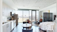This thoughtfully designed single-floor studio offers a seamless blend of contemporary urban living and curated artistry. The space is entered through a striking gallery-style hallway, where crisp white walls serve as a backdrop for a collection of large, black-framed monochromatic artworks and sculptural accents, all set above rich hardwood flooring and accented by a decorative area rug.

The open-plan main living area is anchored by dramatic floor-to-ceiling glass walls that frame expansive city skyline views, flooding the interior with abundant natural light. The exposed concrete ceiling lends an industrial-chic quality to the space, complementing the warm hardwood floors throughout. A dedicated sitting area with leather accent chairs and a round coffee table transitions naturally into a sleeping zone, subtly divided by a slatted room divider for a sense of privacy without sacrificing openness.

The kitchen is efficiently designed with light wood cabinetry, white quartz countertops, a decorative tile backsplash, and a full suite of stainless steel appliances, including a range, dishwasher, and over-the-range microwave. A compact dining area sits just adjacent, ideal for everyday meals.

The bathroom features a clean, modern vanity with wood-toned cabinetry, white countertops, and tile flooring, opening to a well-organized hallway with built-in shelving and louvered closet doors, completing this polished urban retreat.