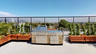 This rooftop terrace is a thoughtfully designed outdoor retreat that combines functionality with an open-air ambiance. The space is anchored by a fully equipped outdoor kitchen, featuring a stainless steel built-in grill with stone accent panels, multiple burners, storage drawers, and ample counter space — ideal for al fresco cooking and entertaining. Flanking the kitchen station are terracotta-toned raised planters that add a warm, organic touch to the modern aesthetic.

The sun deck area offers generous open space furnished with sleek white chaise lounges and side tables, perfect for relaxing under the expansive blue sky. Lush greenery lines the perimeter, with tall, well-maintained shrubs and potted plants creating a sense of privacy and a vibrant natural backdrop throughout the terrace.

A dark metal railing borders the rooftop, providing unobstructed panoramic views of the surrounding cityscape, treetops, and distant rooftops. The clean, light-toned flooring enhances the brightness of the space, reflecting natural sunlight throughout the day. The overall atmosphere is bright, airy, and inviting — a serene elevated escape well-suited for both social gatherings and quiet outdoor relaxation.