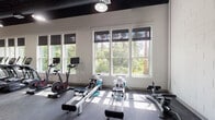 Located at 9010 Running Bull Rd in Orlando, FL, this single-floor fitness center is thoughtfully designed to accommodate a wide range of workout styles and fitness goals. The space features a striking industrial aesthetic, with an exposed black ceiling that contrasts sharply against white painted cinder block walls, creating a modern and energetic atmosphere throughout.

The cardio section is lined with large windows fitted with roller shades, offering abundant natural light and lush views of the surrounding tree canopy. This area houses a row of treadmills, stationary bikes — including a Peloton — and multiple water rowing machines, allowing members to train with both variety and scenic backdrop.

The strength training zone is anchored by a comprehensive selection of Hoist selectorized machines and a full dumbbell rack, providing options for targeted muscle training. Pendant lighting illuminates this section, complementing the open sightlines across the floor.

A standout feature is the dedicated functional training zone, surfaced with artificial turf and equipped with a multi-functional training rig featuring battle ropes, medicine balls, and pull-up bars. A large tractor tire adds to the functional fitness offerings, while open floor space nearby supports group exercises or stretching. A hardwood-floored area rounds out the facility, ideal for yoga, stretching, or low-impact activities.