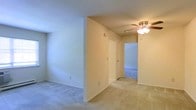 This single-floor residential space is designed with a straightforward, functional layout that prioritizes comfort and ease of movement. The interior is finished with soft, neutral-toned walls that create a clean and welcoming atmosphere throughout. Wall-to-wall carpeting in a light beige hue runs consistently across the main living areas, adding warmth underfoot and contributing to the overall cohesive aesthetic.

The open layout allows for a smooth visual flow between spaces, with a partial wall dividing areas while maintaining a sense of openness. A ceiling fan with an integrated light fixture is centrally positioned, providing both air circulation and ambient lighting to the space.

A hallway connects the main living area to additional rooms, offering a glimpse of further spaces beyond, including what appears to be a room with distinct flooring. A closet door is visible along the corridor, providing convenient built-in storage. Clean white baseboards and door trim add a crisp finishing detail throughout.

The overall ambiance is calm and neutral, making the space highly adaptable for a variety of furnishing styles and personal preferences. The single-floor layout ensures easy accessibility, and the connected room configuration supports a practical and efficient flow throughout the home.