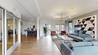 This single-floor leasing and community space for ABQ Uptown offers a thoughtfully designed, open-plan interior finished with warm hardwood-style flooring, white walls, and crisp architectural details throughout. The layout is divided into two primary zones that flow seamlessly into one another.

The main lounge area is anchored by a striking geometric wall mural and a modern brass Sputnik chandelier, creating a bold focal point. Comfortable gray sofas and mid-century style leather accent chairs are arranged around a white marble-top coffee table, while large floor-to-ceiling glass doors and windows flood the space with abundant natural light and provide views of the surrounding streetscape.

Adjacent to the lounge, a bar-height seating area with dark stools sits alongside a compact refreshment counter, offering a casual gathering space. A dining-style table with chairs and vibrant botanical artwork adds color and warmth to this zone.

The office and administrative section features a sleek white reception desk beneath a curved architectural ceiling detail, alongside glass-walled private meeting rooms that maintain an open, transparent feel. A dark wood credenza with botanical artwork and a fully equipped private office complete the professional amenities. Lush potted plants are positioned throughout, enhancing the fresh, welcoming atmosphere of the entire space.