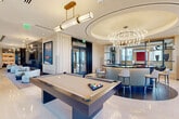 Located in the heart of Fort Lauderdale at 201 SE Sixth Ave, this sophisticated amenity floor offers a generously scaled, single-level space defined by upscale contemporary design and an abundance of natural light. Polished marble-tiled floors and warm wood-paneled walls set an elegant tone throughout, while floor-to-ceiling windows frame sweeping panoramic views of the Fort Lauderdale skyline and surrounding landscape, including swaying palm trees and open sky.

The space is organized into several distinct yet interconnected zones. A sleek entertainment area anchors one section, featuring a full-size billiards table beneath a pair of modern gold pendant lights and a coffered ceiling detail. Adjacent to it, a showstopping circular wet bar commands attention with its fluted wood base, dark marble countertop, and an eye-catching sculptural chandelier above, all framed by expansive glass windows with outdoor views.

A formal dining area offers a long statement table with plush sage-green velvet chairs beneath a cascading crystal chandelier, ideal for private gatherings. Separate lounge areas feature curved sofas, sculptural accent chairs, and a dramatic dark marble fireplace wall with integrated display shelving and large-scale artwork. A dedicated conference-style meeting room rounds out the offering, providing a refined setting for professional engagements within this world-class amenity space.