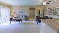 This single-floor residence showcases an open-concept layout that seamlessly connects the living, dining, and kitchen areas into one cohesive and functional space. The living area is centered around plush, light-toned carpeting and features a comfortable gray sofa accented with decorative pillows, complemented by a dark wood coffee table and framed wall art that adds a refined touch. Warm beige walls flow throughout, creating a calm and inviting ambiance.

A distinctive arched entryway serves as an architectural focal point, adding a touch of elegance to the open floor plan. A ceiling fan provides both style and practicality overhead. The dining area is positioned adjacent to the living space and features dark wood chairs arranged around a table, accompanied by a decorative mirror and floral arrangement that enhance the area's visual appeal.

The kitchen transitions naturally into the shared living space, partially visible with a granite-style countertop edge suggesting quality finishes. Neutral tones, consistent flooring materials, and thoughtful decorative elements throughout create a harmonious, well-balanced interior that is both comfortable and polished.