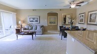 This single-floor residence showcases an open-concept layout that seamlessly connects the living, dining, and kitchen areas into one cohesive and functional space. The living area is centered around plush, light-toned carpeting and features a comfortable gray sofa accented with decorative pillows, complemented by a dark wood coffee table and framed wall art that adds a refined touch. Warm beige walls flow throughout, creating a calm and inviting ambiance.

A distinctive arched entryway serves as an architectural focal point, adding a touch of elegance to the open floor plan. A ceiling fan provides both style and practicality overhead. The dining area is positioned adjacent to the living space and features dark wood chairs arranged around a table, accompanied by a decorative mirror and floral arrangement that enhance the area's visual appeal.

The kitchen transitions naturally into the shared living space, partially visible with a granite-style countertop edge suggesting quality finishes. Neutral tones, consistent flooring materials, and thoughtful decorative elements throughout create a harmonious, well-balanced interior that is both comfortable and polished.