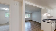 Located at 8106 SE Lake Rd in Portland, Oregon, this one-bedroom, one-bathroom apartment offers a functional and efficient single-floor layout. The living room is bright and welcoming, featuring light hardwood-style flooring, a textured ceiling, and a large picture window dressed with vertical blinds that frames lush outdoor greenery, filling the space with natural light. Baseboard heating runs beneath the window, providing consistent warmth throughout. The galley-style kitchen flows directly from the living area and is equipped with white cabinetry, dark countertops, a double sink, a white electric range, and a refrigerator, all set atop matching hardwood-style flooring. The private bedroom offers plush carpeting, a window with outdoor views, and baseboard heating, creating a cozy and comfortable retreat. An electrical panel is conveniently accessible within the unit. The full bathroom includes a clean, white tub-shower combination with a curved curtain rod and standard fixtures. Throughout the unit, neutral white walls and clean finishes contribute to a bright, understated aesthetic that provides a versatile canvas for personalization.