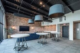 This single-floor space in Birmingham, Alabama, masterfully combines industrial heritage with contemporary design. The defining architectural feature throughout is the original exposed brick, which lines nearly every wall and lends warmth and texture to the open interior. Large-format gray tile flooring flows consistently across the main areas, grounding the space with a clean, modern contrast to the rustic brickwork above.

The main gathering area is spacious and open, accommodating multiple seating configurations. A lounge zone features deep blue tufted sofas and ottomans arranged around a wall-mounted flat-screen television, flanked by pendant lighting and potted olive trees that soften the industrial aesthetic. Overhead, exposed ductwork and black ceiling infrastructure reinforce the building's raw character.

Floor-to-ceiling steel-framed glass panels open toward an outdoor terrace, filling the interior with natural light and providing seamless indoor-outdoor connectivity. String lights visible through the glass hint at the terrace's entertaining potential.

A dedicated kitchenette area features warm wood cabinetry, white countertops, and exposed brick walls, complemented by a water station and commercial-style glass-door refrigerator nearby. An elevator provides additional accessibility. The overall ambiance is refined yet relaxed, well-suited for events, gatherings, or collaborative use.