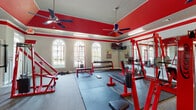 Located at 800 Gunn Rd in Centerville, Georgia, this single-floor fitness facility is designed to deliver a comprehensive workout experience in a visually striking environment. The space is defined by its bold red-painted upper walls and ceiling trim contrasted against light gray lower walls, creating an energetic and motivating atmosphere throughout.

The facility is generously sized, offering ample floor space across distinct training zones. A dedicated free weights area features a full dumbbell rack, flat benches, and rubber-matted flooring ideal for lifting. Adjacent to this, a strength training zone houses multiple red-framed Legend brand cable machines, squat racks, and press stations, providing a complete range of resistance training options.

The cardio section includes multiple treadmills equipped with digital displays, along with stationary cycling equipment. Full-length mirrors line the walls, enhancing the sense of space and allowing users to monitor their form during exercise.

Architecturally, the space features vaulted ceilings, elegant arched windows with decorative transoms that flood the interior with natural light, and multiple ceiling fans that promote airflow. Motivational artwork and signage decorate the walls, adding character to the space. The combination of durable rubber and carpet flooring throughout accommodates both heavy equipment and high-traffic use with ease.