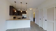 This single-floor space showcases a thoughtfully designed open-concept layout, centered around a contemporary kitchen that serves as the heart of the home. The kitchen features dark espresso cabinetry that contrasts elegantly with light granite countertops on the extended island, which includes a built-in sink and offers additional workspace or casual seating. A subway tile backsplash adds texture and visual interest to the cooking area, while stainless steel appliances — including a French door refrigerator with an ice and water dispenser, a smooth-top range, and an over-the-range microwave — complete the modern aesthetic. Three Edison-style pendant lights hang above the island, complementing the track lighting overhead to create a warm, well-lit atmosphere. Light-toned wood-look flooring flows throughout the space, enhancing the bright and airy feel created by the white walls and ceilings. A hallway visible from the kitchen leads to additional rooms with white paneled doors, suggesting a well-organized and functional floor plan. The overall ambiance is clean, contemporary, and move-in ready.