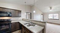 Located at 1740 S Katie Ave in Sioux Falls, South Dakota, this single-floor unit offers a thoughtfully designed layout with consistent finishes throughout. The kitchen serves as a focal point, featuring dark espresso cabinetry, granite-style countertops, and black appliances including a refrigerator, electric range, and built-in microwave. A double-basin stainless steel sink sits along an extended peninsula that opens toward the living area, enhanced by vaulted ceilings and pendant lighting that lend an open, airy quality to the space. Stone-look tile flooring grounds the kitchen and carries into the bathroom areas.

The bedrooms are finished with neutral carpet and receive natural light through sliding windows that offer views of the surrounding neighborhood. Dark-trimmed door frames and baseboards provide a clean contrast against the light-colored walls throughout. The carpeted hallway connects the living spaces to the private areas of the unit seamlessly.

The bathroom is well-equipped with a full tub-shower combination, a dark wood vanity topped with granite-style counters, a framed mirror, and stone-look tile flooring that mirrors the kitchen. The overall ambiance is clean, neutral, and move-in ready, with cohesive design elements tying each area of the unit together.