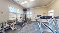 This fitness center offers a comprehensive workout environment designed to support a wide range of exercise routines. The space features elevated ceilings accented by elegant crown molding, contributing to an open and airy atmosphere. Multiple windows fitted with plantation shutters line the walls, allowing generous natural light to fill the room while providing adjustable privacy.

The flooring consists of patterned carpet tiles in neutral blue and gold tones, with protective rubber mats placed beneath the exercise equipment. The facility is equipped with a variety of cardio machines, including a treadmill and a recumbent stationary bike, as well as multiple cable and weight-stack strength machines suitable for both upper and lower body training. A dedicated free weight area includes a dumbbell rack and a flat workout bench, catering to those who prefer traditional resistance training.

Multiple wall-mounted flat-screen televisions are positioned throughout the space, including one suspended from the ceiling for broader visibility, ensuring an engaging workout experience from any angle. A wall-mounted accessory station provides sanitation supplies, reinforcing cleanliness standards. A doorway connects to an adjacent room, adding functional utility. The overall design is clean, well-organized, and purposefully laid out to maximize usability within its footprint.