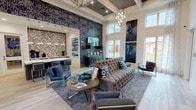 This leasing office is a thoughtfully designed, single-floor space that blends professional functionality with upscale interior design. The main lounge area commands attention with soaring double-height ceilings, floor-to-ceiling windows that flood the space with natural light, and sweeping views of the surrounding grounds. A tufted velvet sofa, sleek modern armchairs, and layered area rugs anchor the seating area, while a dramatic patterned feature wall and bold curated artwork in a navy and dark blue palette establish a refined aesthetic throughout. Pendant chandeliers and recessed lighting complement the coffered ceiling details, adding warmth and dimension to the space. Light hardwood-style flooring runs continuously throughout, enhancing the open and airy feel. The kitchenette area is well-appointed with crisp white quartz countertops, taupe cabinetry, a stainless steel sink, and a striking herringbone tile accent wall that serves as a focal point. Pendant lighting in brushed nickel adds a modern touch, and the space is stocked for guest hospitality. A dedicated private office provides a quiet, professional environment for one-on-one consultations, featuring a dark wood desk, guest seating, and tasteful artwork. Together, the spaces create a welcoming and polished environment that effectively communicates the quality and character of the community it represents.