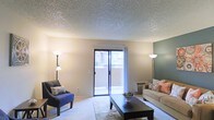 This single-floor space offers a comfortable and well-appointed layout designed for easy, everyday living. The living area serves as the heart of the space, featuring plush wall-to-wall carpeting that adds warmth underfoot and a classic textured ceiling overhead. Neutral beige walls are complemented by an accent wall in a muted sage green, lending a subtle pop of color and visual depth to the room. Natural light pours in through a large sliding glass door, which opens to an outdoor balcony and frames views of the surrounding greenery, creating a seamless connection between the interior and exterior. The sliding door is flanked by a traditional window, further enhancing the brightness of the space. A wall-mounted HVAC unit ensures year-round comfort. Floor lamps positioned on either side of the room provide warm ambient lighting for the evenings. The overall ambiance is inviting and relaxed, with a clean, understated aesthetic that offers a versatile backdrop for a variety of interior styles. The single-floor layout provides a practical and accessible flow throughout the space, making it functional and easy to navigate.