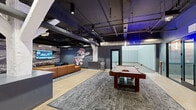 Located at 321 Elm St in Cincinnati's 45202 zip code, this amenity floor at Textile Apartments delivers an impressive range of recreational and social spaces. The centerpiece of the lounge area is a full-size pool table set atop layered area rugs, surrounded by tan leather sofas and complemented by a large-screen TV mounted against a dramatic black-and-white stadium mural — a clear nod to Cincinnati's baseball heritage, complete with a bold Mr. Redlegs mascot graphic. Exposed ductwork and an open ceiling grid painted dark give the space an industrial-chic character, while light wood-plank flooring adds warmth throughout. A glowing "GAME ON" neon sign and blue ambient lighting set an energetic, social atmosphere. The lounge flows into a flexible dining and seating zone outfitted with mixed-material tables and modern chairs, ideal for casual gatherings. A wide corridor with banquette seating and eclectic wall art connects the main lounge to additional spaces. The crown jewel of the amenity level is a full indoor basketball half-court, featuring a hardwood sports floor with regulation markings, a wall-mounted glass backboard, and padded wall protection — all housed beneath soaring ceilings with bright overhead lighting.