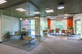 Located at 800 W Marietta St NW in Atlanta, Georgia, this thoughtfully designed single-floor office space blends functionality with a vibrant, modern aesthetic. The layout promotes collaboration through a variety of distinct work zones, each carefully furnished to support different working styles.

At the heart of the space is a generous open work area featuring long, light-toned communal tables paired with ergonomic chairs in blue and white, ideal for team collaboration or focused individual work. A warm-toned wood built-in bench runs along the windowed wall, accented by pendant lighting and lush greenery, offering a more relaxed seating option with views of the surrounding tree canopy.

Glass-walled enclosures provide semi-private meeting areas without sacrificing the open, airy feel of the space. One enclosed room features a round table with modern chairs and a sculptural arc lamp, while a larger adjacent conference area accommodates groups with a full set of colorful seating. Bold orange accent walls and a distinctive illustrated wallpaper add personality and visual interest throughout.

Acoustic tile ceilings with flush and pendant lighting ensure a well-lit, comfortable environment. Natural light floods in through expansive windows along multiple walls, maintaining a strong connection to the outdoors. The overall ambiance is energetic yet focused, making this office an inspiring setting for creative and professional work alike.